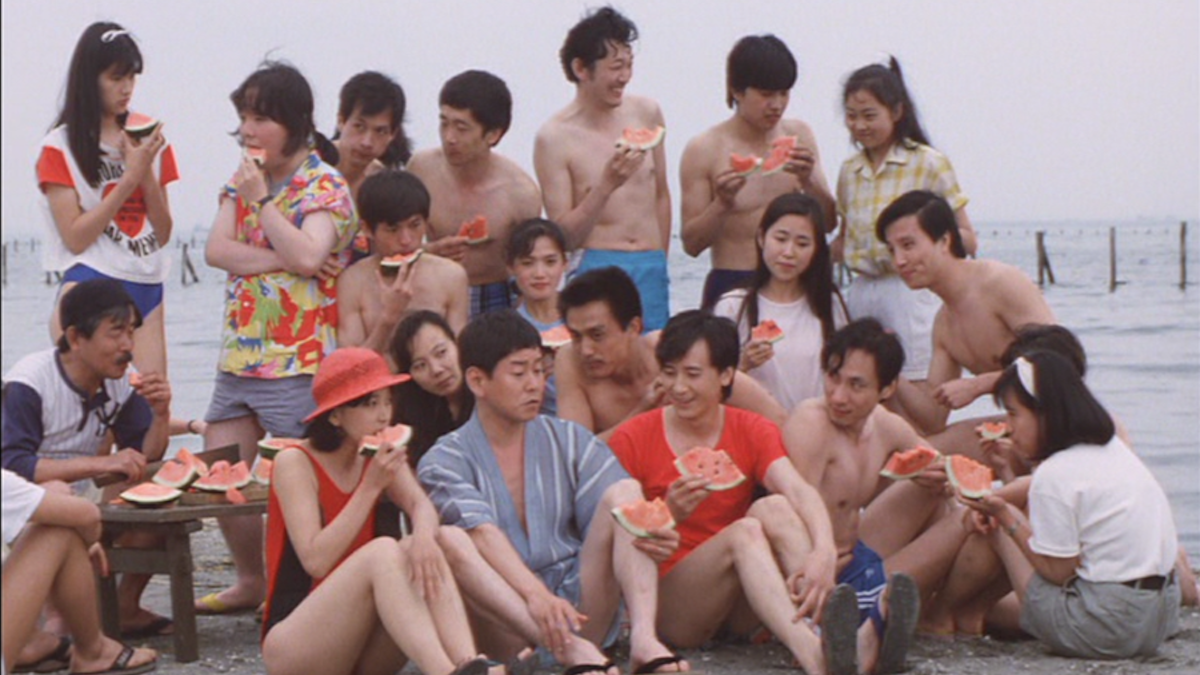 large group of people sitting in front of water eating watermelon